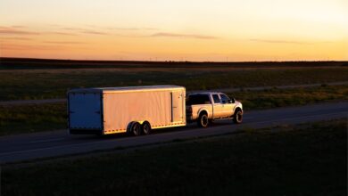 A truck is hauling a trailer down a road. The sun is low on the horizon, causing lowlight conditions.