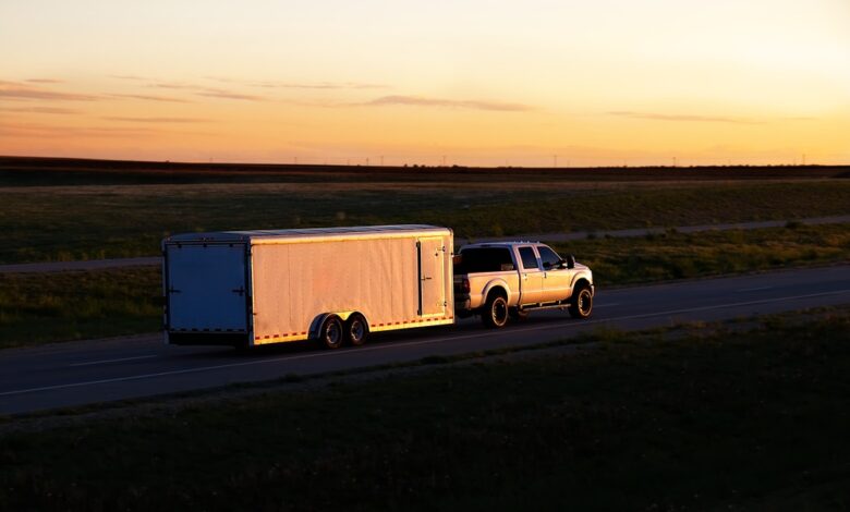 A truck is hauling a trailer down a road. The sun is low on the horizon, causing lowlight conditions.