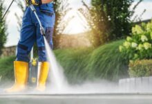 A man is standing while using a power washer on a surface. The man is wearing blue pants.