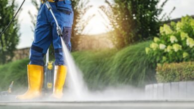 A man is standing while using a power washer on a surface. The man is wearing blue pants.