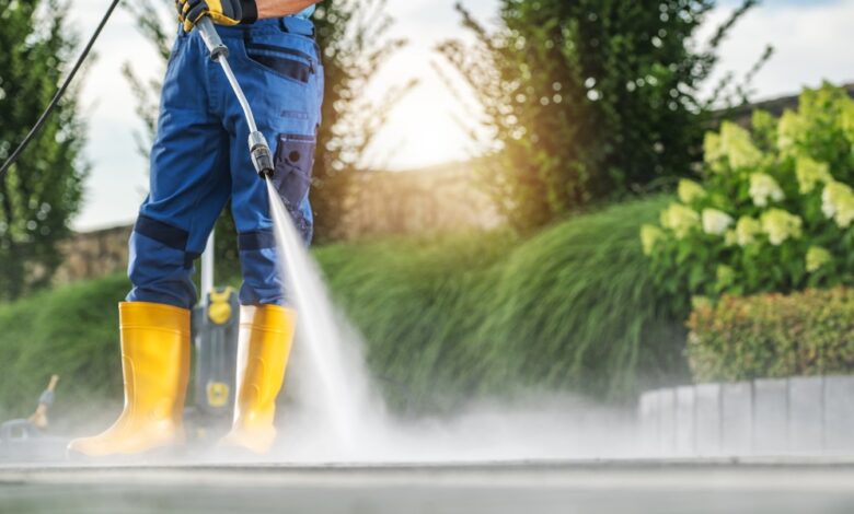 A man is standing while using a power washer on a surface. The man is wearing blue pants.