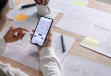  Person tests a smartphone interface at a desk covered with hand-drawn app wireframes, sticky notes, and pens.