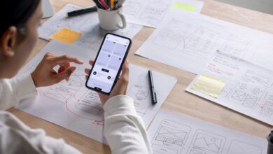  Person tests a smartphone interface at a desk covered with hand-drawn app wireframes, sticky notes, and pens.