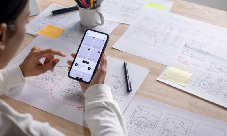  Person tests a smartphone interface at a desk covered with hand-drawn app wireframes, sticky notes, and pens.