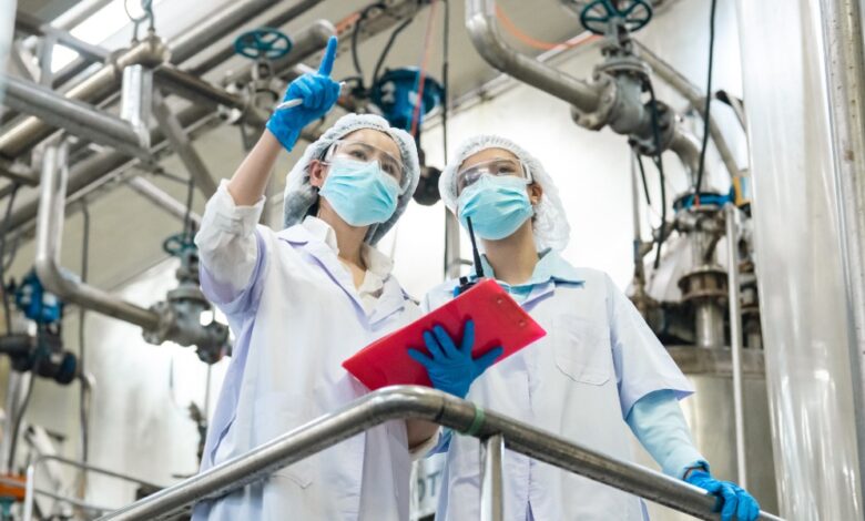 Two workers in masks, gloves, and white coats inspect industrial piping while one points and the other holds a tablet.
