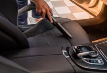 A man is using a vacuum to clean the driver’s seat of a car. The seat is black fabric, and the vacuum is black plastic.