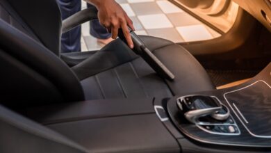 A man is using a vacuum to clean the driver’s seat of a car. The seat is black fabric, and the vacuum is black plastic.