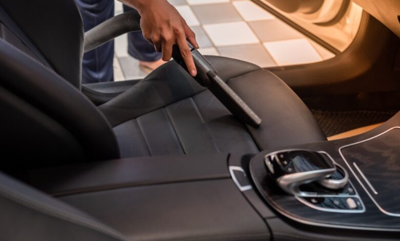 A man is using a vacuum to clean the driver’s seat of a car. The seat is black fabric, and the vacuum is black plastic.