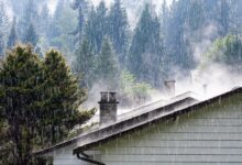 Strong rain falls on a house roof with steam rising near chimneys and tall evergreen trees in the background.