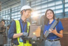A man in a high-visibility vest and a hard hat holding a laptop smiles next to a woman holding a part in a factory.