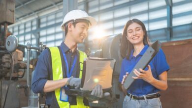 A man in a high-visibility vest and a hard hat holding a laptop smiles next to a woman holding a part in a factory.