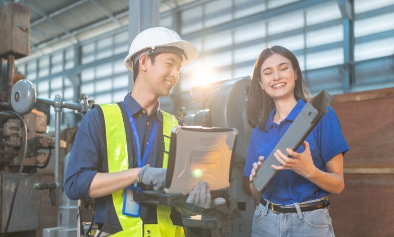 A man in a high-visibility vest and a hard hat holding a laptop smiles next to a woman holding a part in a factory.