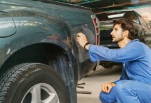 A man in a blue jumpsuit is kneeling down next to a truck. The man is in a garage environment.