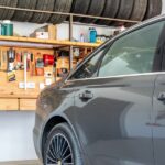 A gray car sits inside a home garage with an organized workbench, hand tools, supplies, and stored tires.
