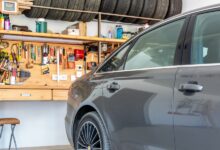 A gray car sits inside a home garage with an organized workbench, hand tools, supplies, and stored tires.