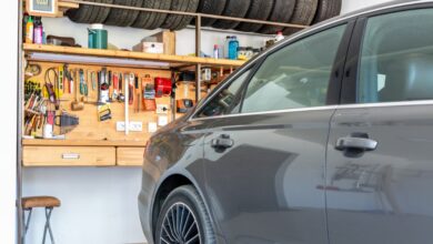 A gray car sits inside a home garage with an organized workbench, hand tools, supplies, and stored tires.