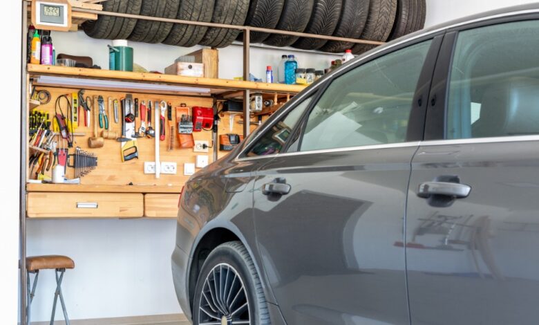 A gray car sits inside a home garage with an organized workbench, hand tools, supplies, and stored tires.