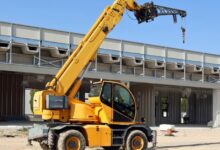 A yellow telehandler parked in the middle of a construction site. The boom arm is extended above the telehandler.