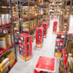 A warehouse has towering industrial shelves holding cardboard boxes. In the aisle, robots move stacks of products.